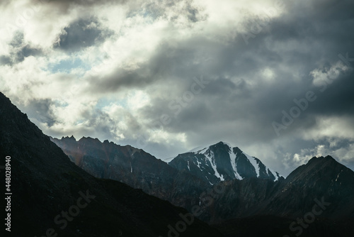 Atmospheric mountain landscape with silhouettes of rocks and snowbound sharp pinnacle under cloudy sky. Great rocks with snow. Beautiful mountain scenery with snow peaked top. Snow-capped pointy peak.