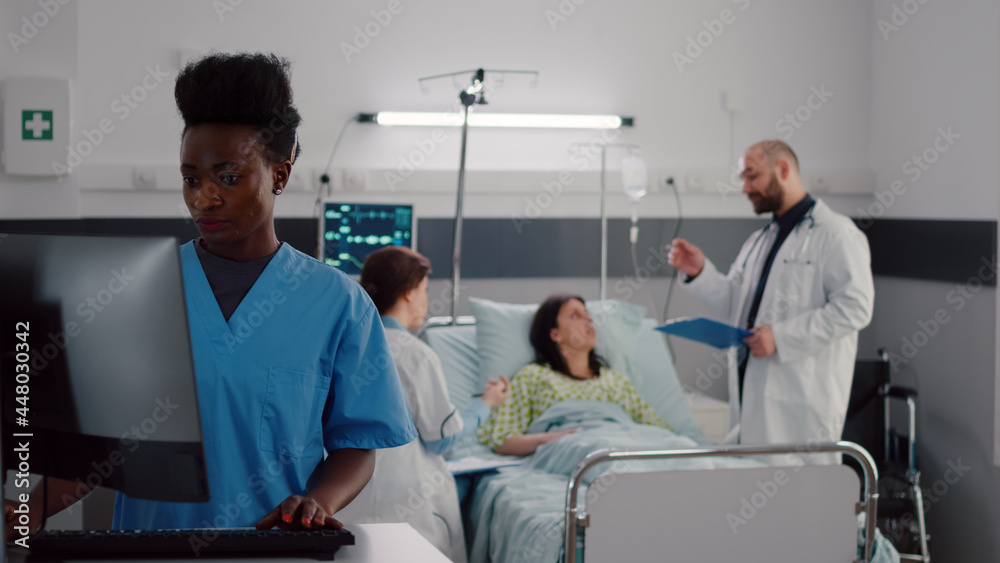 Obraz premium Black nurse typing disease symptom while specialist practitioner doctors checking sick woman writing health treatment on clipboard during medical appointment. Patient resting in bed in hospital ward