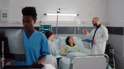 Black nurse typing disease symptom while specialist practitioner doctors checking sick woman writing health treatment on clipboard during medical appointment. Patient resting in bed in hospital ward