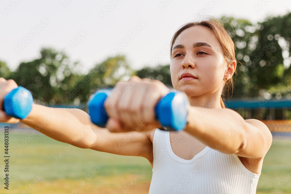 Fitness girl workout with dumbbells, sporty woman in sportswear doing intense exercise outdoor in stadium, training for strong muscular body, wearing white top. Fitness girl workout with dumbbells, sporty woman in sportswear doing intense exercise outdoor in stadium, training for strong muscular body, wearing white top.
