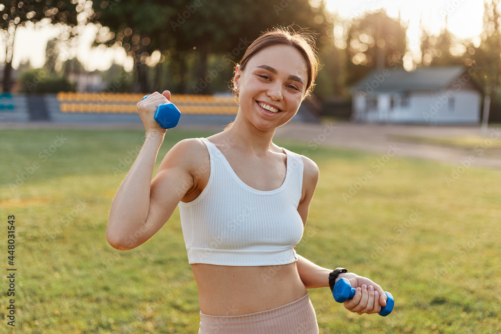Portrait of young adult attractive smiling woman, being happy to workout in stadium outdoor, holding blue dumbbells in hands, looking at camera. Portrait of young adult attractive smiling woman, being happy to workout in stadium outdoor, holding blue dumbbells in hands, looking at camera.