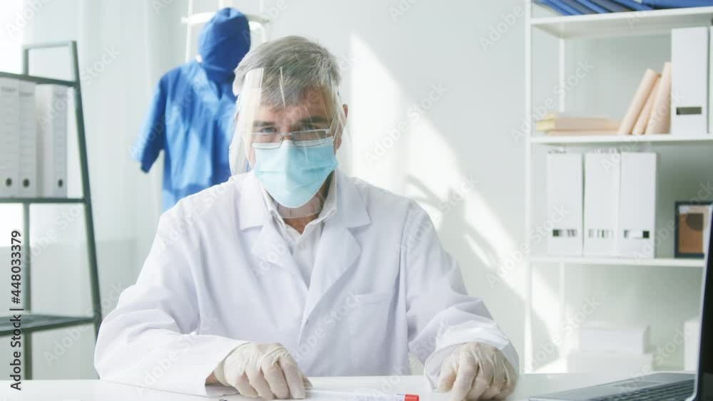 PCR test in laboratory, aged doctor in glasses holding COVID-19 swab collection kit and looking in camera, wearing PPE medical suit, mask, gloves, test tube for taking OP NP patient specimen sample PCR test in laboratory, aged doctor in glasses holding COVID-19 swab collection kit and looking in camera, wearing PPE medical suit, mask, gloves, test tube for taking OP NP patient specimen sample