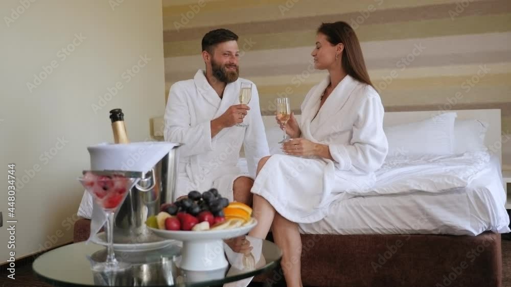 Couple in white coats are sitting on a bed in a hotel with glasses of champagne. Couple in white coats are sitting on a bed in a hotel with glasses of champagne.