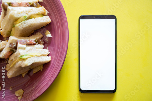 Mobile phone with white screen next to a plate with a sandwich on a yellow table, top view.