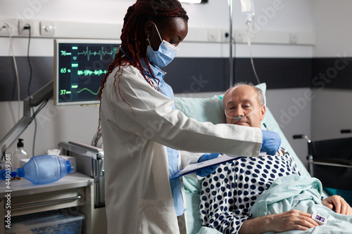 African american medical practitoner wearing chirurgical mask, in hospital room during consultation of sick unwell senior man, breathing with help from test tube and iv drip attached. photo
