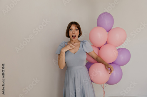Festive portrait of young lady against light background. Handsome girl with smooth dark hair, blue dress and pale skin, surprisingly looking into camera and holding violet and pink balloons