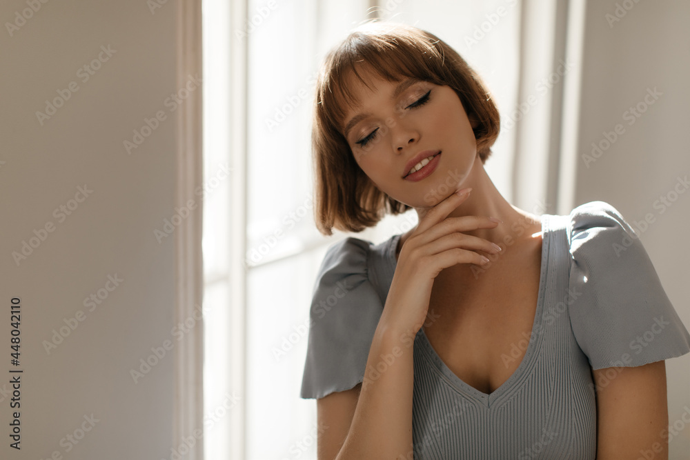 Frontal portrait of young lady against window background. Appealing dark-haired girl with closed eyes, trendy makeup, blue top, touching neck and posing in light room Frontal portrait of young lady against window background. Appealing dark-haired girl with closed eyes, trendy makeup, blue top, touching neck and posing in light room
