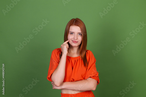 Young red hair woman in casual orange blouse on green background thoughtful thinking