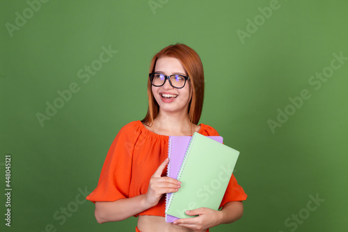 Young red hair woman in casual orange blouse and eye glasses on green background holding notebooks happy excited cheerful