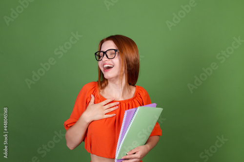 Young red hair woman in casual orange blouse and eye glasses on green background holding notebooks happy excited cheerful laugh