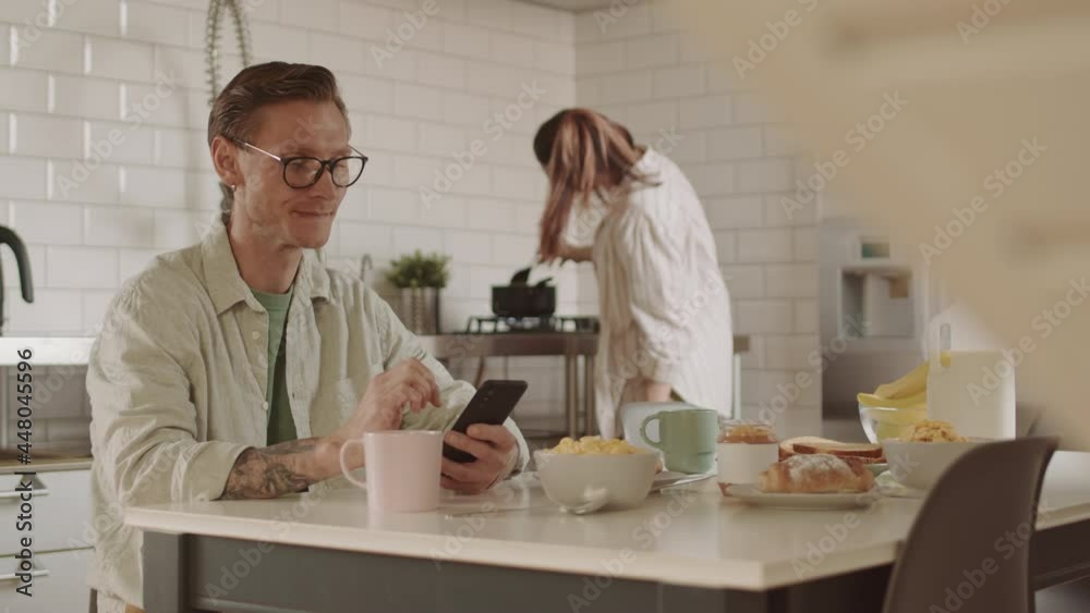 Locked-down of Caucasian man dressed casually wearing glasses, sitting at dining table with breakfast on, using smartphone, talking to wife cooking on background Locked-down of Caucasian man dressed casually wearing glasses, sitting at dining table with breakfast on, using smartphone, talking to wife cooking on background