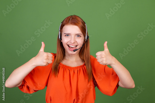 Young red hair woman in casual orange blouse on green background manager call centre help line worker smile shows thumbs up