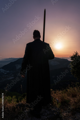 silhouette of a warrior in a leather trench coat with a greatsword at sunset with mountains 