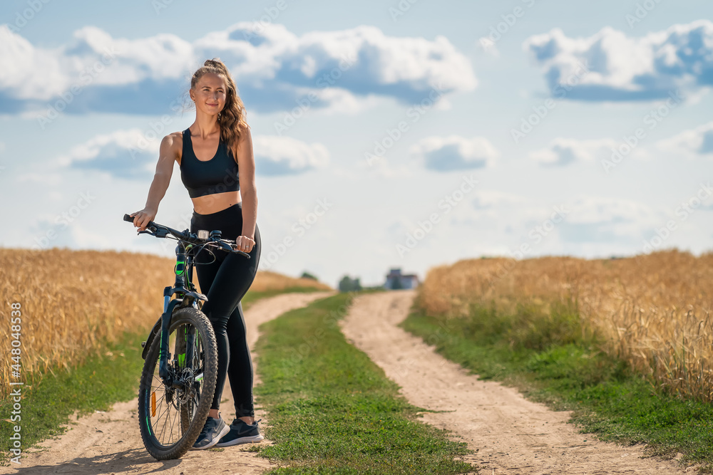 Young sporty cheerful woman in leggings and a top with a bicycle stands on the road in the middle of a field with triticale on a sunny summer day. Concept of a healthy lifestyle Young sporty cheerful woman in leggings and a top with a bicycle stands on the road in the middle of a field with triticale on a sunny summer day. Concept of a healthy lifestyle