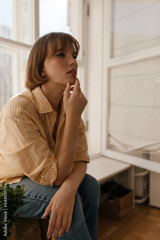 Thoughtful young girl sitting on windowsill of light room. Gorgeous dark-haired lady wearing orange shirt and blue pants, touching chin and looking aside Thoughtful young girl sitting on windowsill of light room. Gorgeous dark-haired lady wearing orange shirt and blue pants, touching chin and looking aside