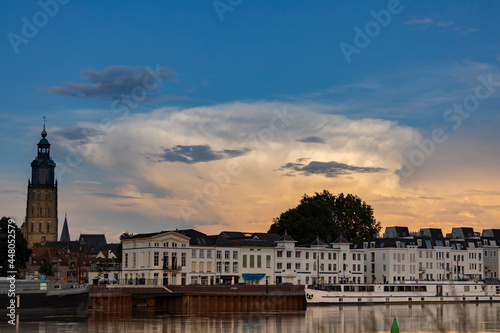 Towering cumulonimbus rain cloud above cityscape of Zutphen in The Netherlands at sunset with orange colors contrasted against a blue sky. Dutch climate and weather condition landscape. © Maarten Zeehandelaar
