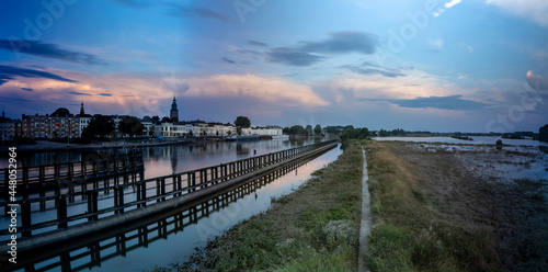 Panorama showing floodplains and cityscape of Zutphen in The Netherlands with large cumulonimbus rain clouds above in colorful sunset tones. Dutch climate weather condition landscape. © Maarten Zeehandelaar