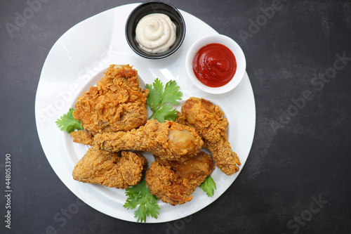 Fresh, hot, crispy deep fried chicken served on a white plate with tomato ketchup and mayonnaise. Chicken snack, hot fried chicken wings. Fast food over moody dark black background with copy space. 
