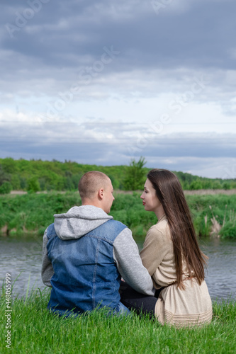 sweet couple sitting by the water river on green grass. attractive brunette woman and bald man outdoors. true love, happy family, romance, successful marriage