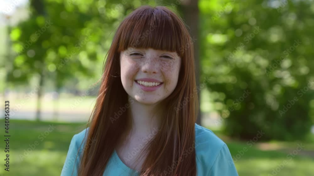 Dolly push shot of smiling redhead teen girl looking at camera standing in summer park Dolly push shot of smiling redhead teen girl looking at camera standing in summer park