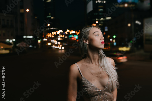 beautiful young blonde long haired woman looking side thoughtfully sad on summer night street in fancy golden cocktail Glided dress. waiting proudly alone. Bokeh lights background.