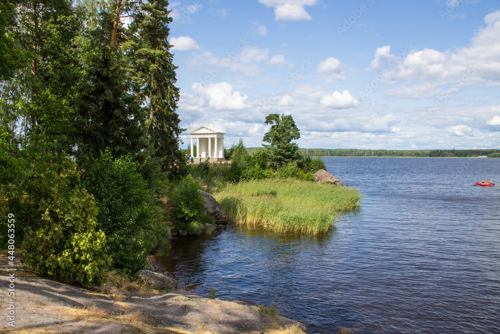Obraz premium VYBORG, Leningrad REGION, RUSSIA-JULY, 19, 2021: view of the white temple of Neptune and the green foliage of trees on a summer cloudy day in the Monrepos Park and the beautiful shore and blue water