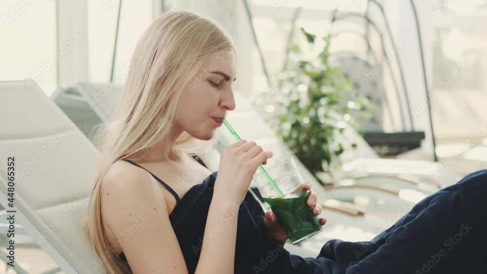 Profile view of blonde young woman drinking cooling mint drink from a straw. She lying on a deck chair and relaxing. Profile view of blonde young woman drinking cooling mint drink from a straw. She lying on a deck chair and relaxing.