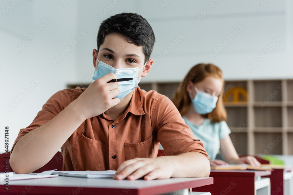 Schoolboy in medical mask holding pen near notebook and blurred friend
