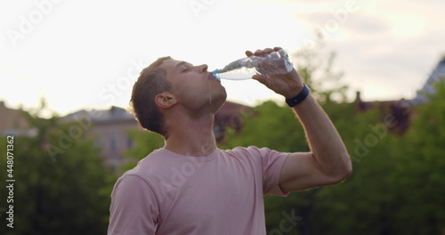 Medium shot of thirsty young man drinking water in plastic bottle outdoors.