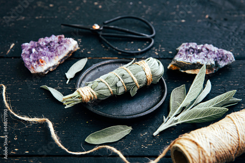 White homemade sage Salvia apiana smudge stick at home with homegrown sage leaves. Cotton string, vintage scissors and amethyst clusters for decoration on black wood background. Side view. photo