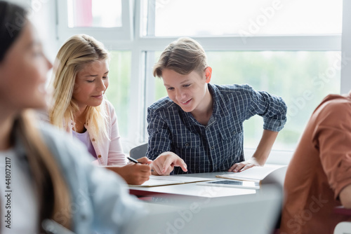 Smiling schoolboy pointing at notebook near friend in classroom