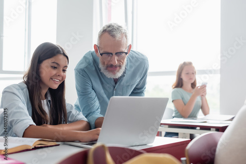 Teacher looking at laptop near smiling schoolgirl in classroom