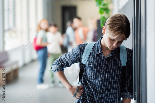 upset schoolboy standing alone with bowed head near blurred pupils in school corridor
