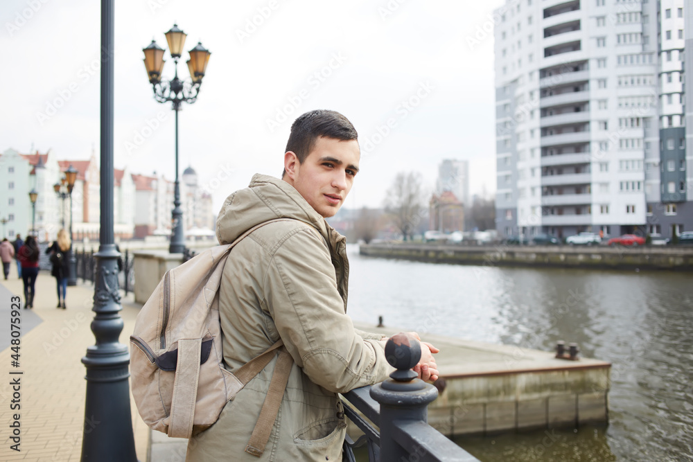 Half length portrait of handsome Caucaisan tourist with casual backpack posing at city embankment, young hipster guy 20s looking at camera during travel vacations for visiting town with quay Half length portrait of handsome Caucaisan tourist with casual backpack posing at city embankment, young hipster guy 20s looking at camera during travel vacations for visiting town with quay