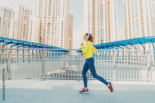 Young woman in bright sportswear quickly runs across a pedestrian bridge in the Dubai Marina district. The concept of a female healthy lifestyle and fitness © EdNurg