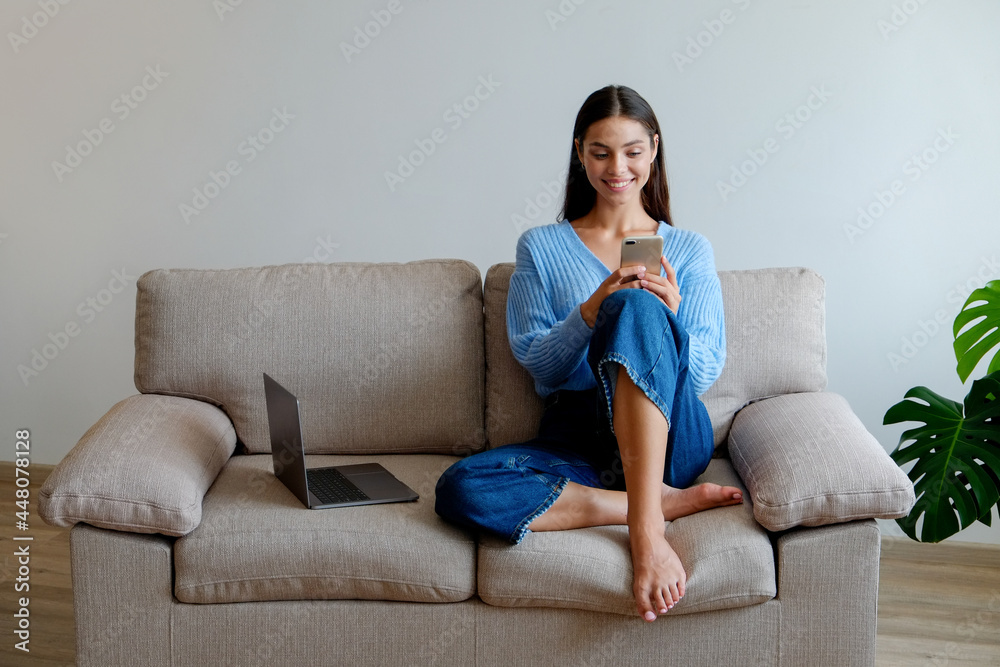 Joyful female having a fun video phone call. Portrait of young brunette woman wearing denim culottes, sitting on the couch texting and smiling. Background, copy space, close up. Joyful female having a fun video phone call. Portrait of young brunette woman wearing denim culottes, sitting on the couch texting and smiling. Background, copy space, close up.