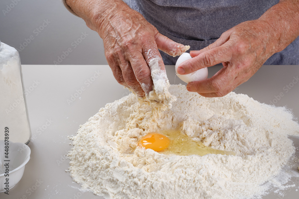 Grandmother breaks an egg for cooking pie. Hand mixing dough on the gray table. Female hands of an elderly woman in a gray apron preparation of dough for a pizza, bread, pasta, festive cake. Grandmother breaks an egg for cooking pie. Hand mixing dough on the gray table. Female hands of an elderly woman in a gray apron preparation of dough for a pizza, bread, pasta, festive cake.