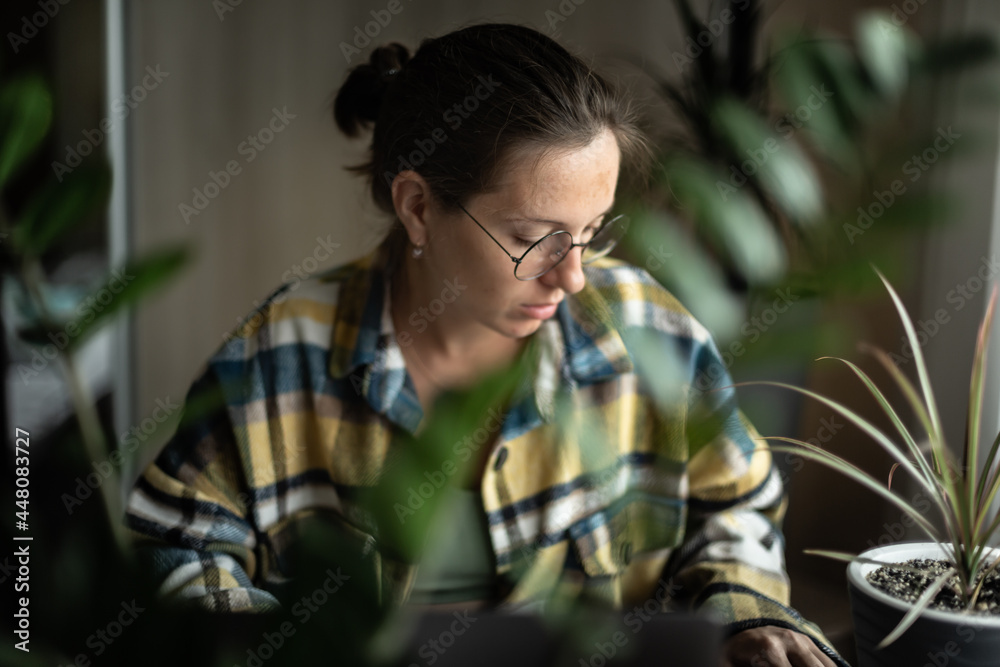 Young woman working on laptop remotely from home, in cozy atmosphere, with flowers on the desk. Pretty female wearing round eyeglasses using laptop