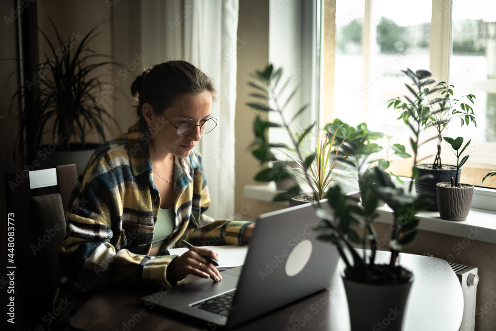 Focused woman in domestic wear and eyeglasses sitting near the window at cozy workplace and browsing laptop while working on startup project remotely at home Focused woman in domestic wear and eyeglasses sitting near the window at cozy workplace and browsing laptop while working on startup project remotely at home