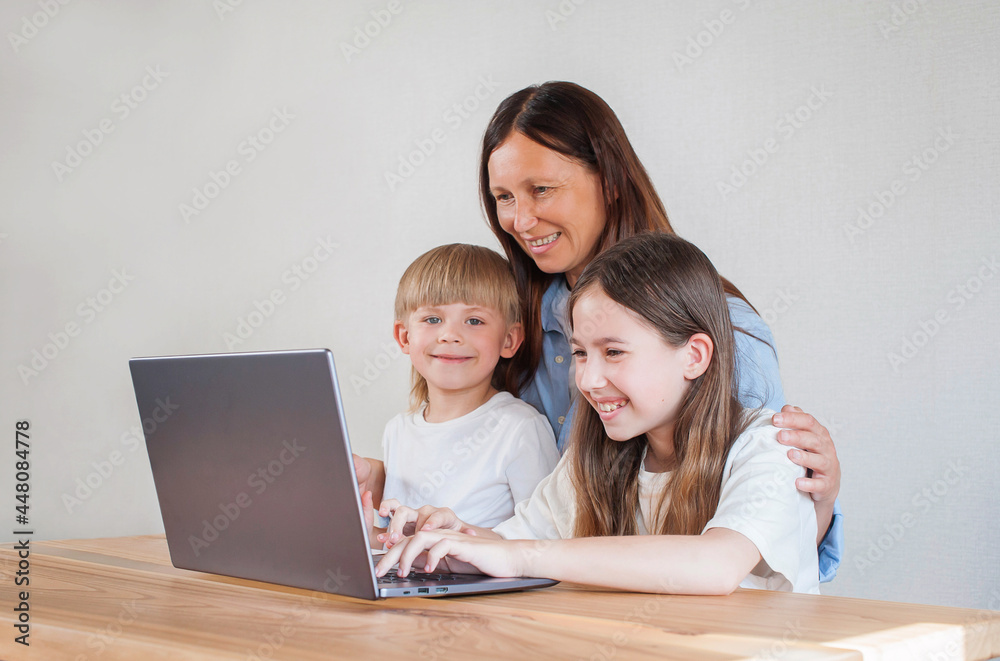 Happy mom with two children are sitting at home at the table with a laptop. Distance learning of children. Mom helps children learn remotely Happy mom with two children are sitting at home at the table with a laptop. Distance learning of children. Mom helps children learn remotely