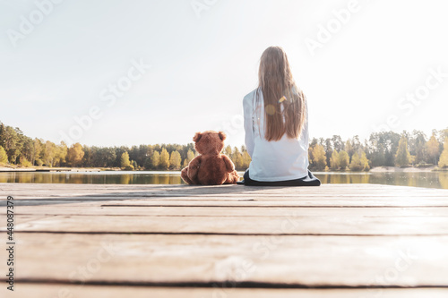 Happy little girl with stuffed toy teddy bear sitting on wooden pier near calm lake against autumn forest on sunny day back view