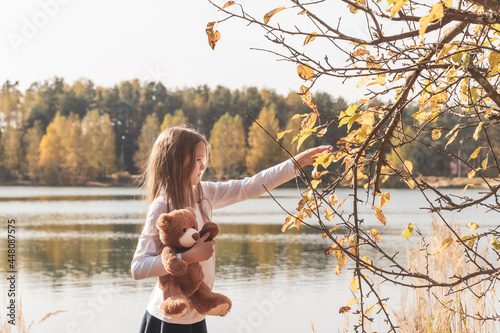 Pretty little girl in school uniform with cute teddy bear holds dry leaf on riverbank in sunny forest on autumn day side view