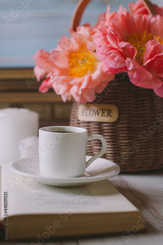 Small coffee cup on an opened book, candles and pink peach peony in basket  on a light blue background, soft selective focus