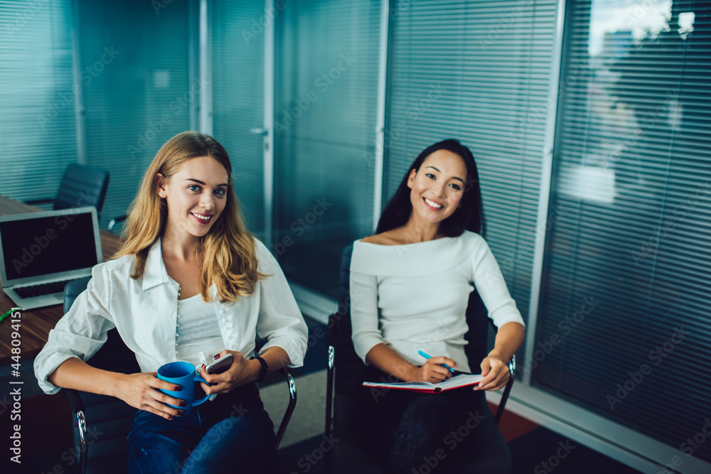 Portrait of successful executive managers dressed in casual clothes smiling at camera during working day in office interior, happy Asian and Caucasian female professionals with textbook posing indoors Portrait of successful executive managers dressed in casual clothes smiling at camera during working day in office interior, happy Asian and Caucasian female professionals with textbook posing indoors