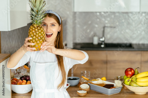 Attractive beautiful woman wearing white shirt, apron holding pineappe and hiding half of her face, looking at camera with happy smile while standing in modern kitchen at home. Healthy food photo