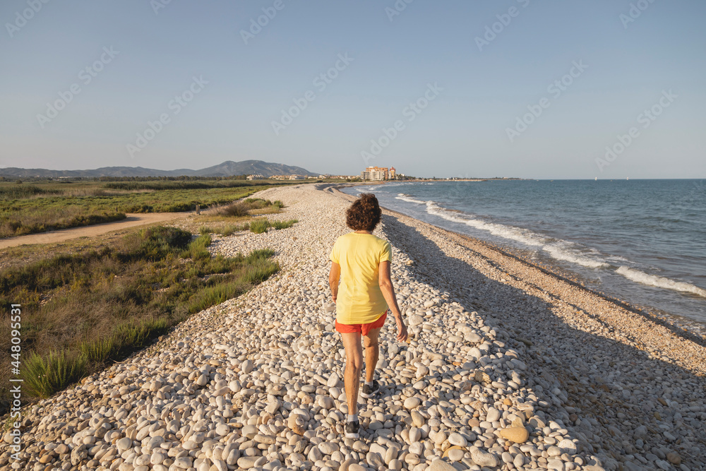 A cheerful and vital woman, in her 70s, walks along a natural pebble beach at sunset, by the Mediterranean Sea, Spain