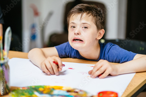 Perception of the world concept. Brunette boy with down syndrome looking at camera sitting at an table with white paper while painting with colorful pencil during painting class.