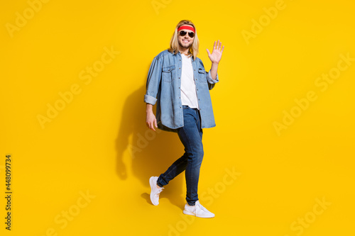 Photo of funky charming young guy dressed denim shirt smiling walking waving arm palm isolated yellow color background