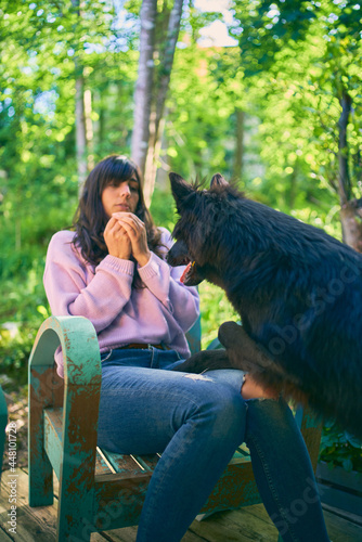 Black dog jumps to young woman's lap, who is wearing jeans and pink sweater and is sitting on a vintage turquoise chair. Focus on the dog.