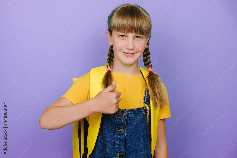 Portrait of beautiful smiling schoolgirl showing thumbs up, happy looking at camera, wears yellow backpack, posing isolated over purple studio background with copy space. Back to school concept Portrait of beautiful smiling schoolgirl showing thumbs up, happy looking at camera, wears yellow backpack, posing isolated over purple studio background with copy space. Back to school concept